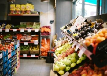 Interior de una frutería o supermercado con estanterías llenas de frutas y verduras frescas, como manzanas, ciruelas, tomates y zanahorias, dispuestas en cajas y bandejas.
